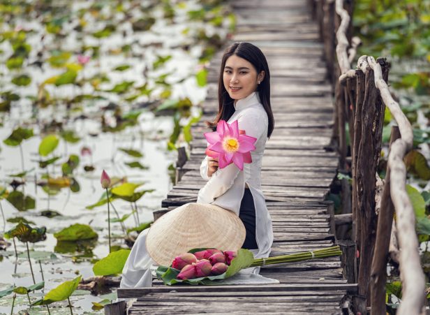 Portrait of beautiful vietnamese woman with traditional vietnam hat holding the pink lotus sitting on the wooden bridge in big lotus lake, vietnam, aisan or southeast asia travel concept